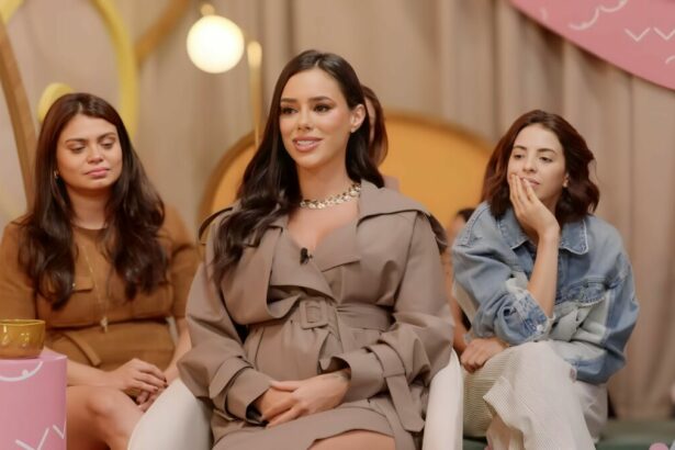 Bruna Biancardi de vestido bege, sorrindo, enquanto fala de Neymar em um podcast; há duas mulheres por trás observando Bruna