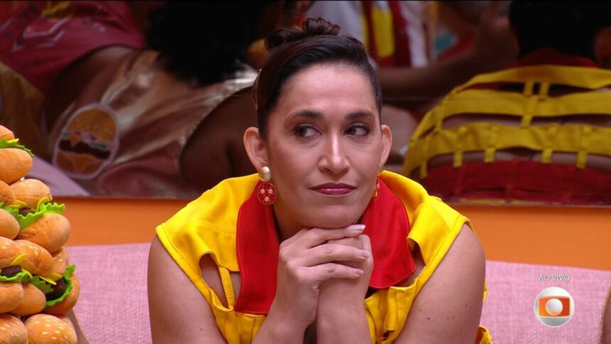Daniele com os braços próximos a boca sentada no sofá da sala do BBB 25 respondendo perguntas de Tadeu