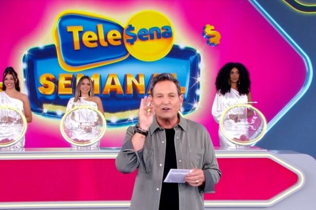 Luis Ricardo levanta a mão para iniciar o sorteio da Tele Sena Semanla edição 49. O cenário está rosa e e ele se posiciona a frente dos globos com as bolas do sorteio