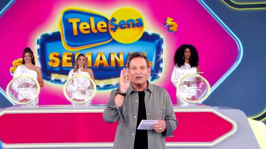 Luis Ricardo levanta a mão para iniciar o sorteio da Tele Sena Semanla edição 49. O cenário está rosa e e ele se posiciona a frente dos globos com as bolas do sorteio