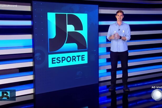 Pedro Ramiro em pé no cenário do Jornal da Record. Ele veste o uniforme esportivo da RECORD e está em frente ao telão do telejornal