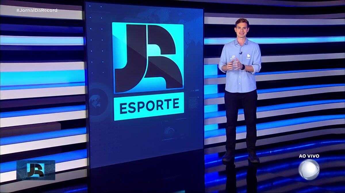 Pedro Ramiro em pé no cenário do Jornal da Record. Ele veste o uniforme esportivo da RECORD e está em frente ao telão do telejornal