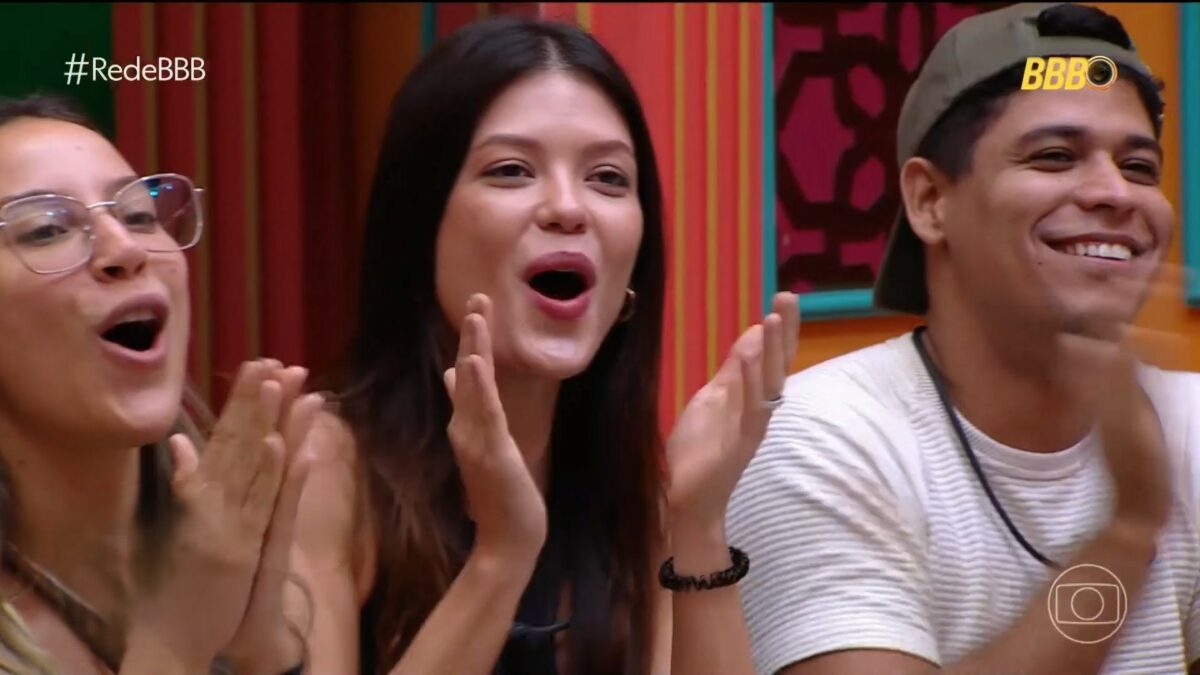 Renata, Vitória Strada e Guilherme batem palma sentados no sofá da sala do BBB 25 ao descobrirem prêmio final
