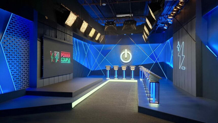 Sala de Apostas do Power Couple Brasil 7 com luzes de neon verde e azul, telão com logo do Power Couple e púlpitos onde os casais fazem apostas