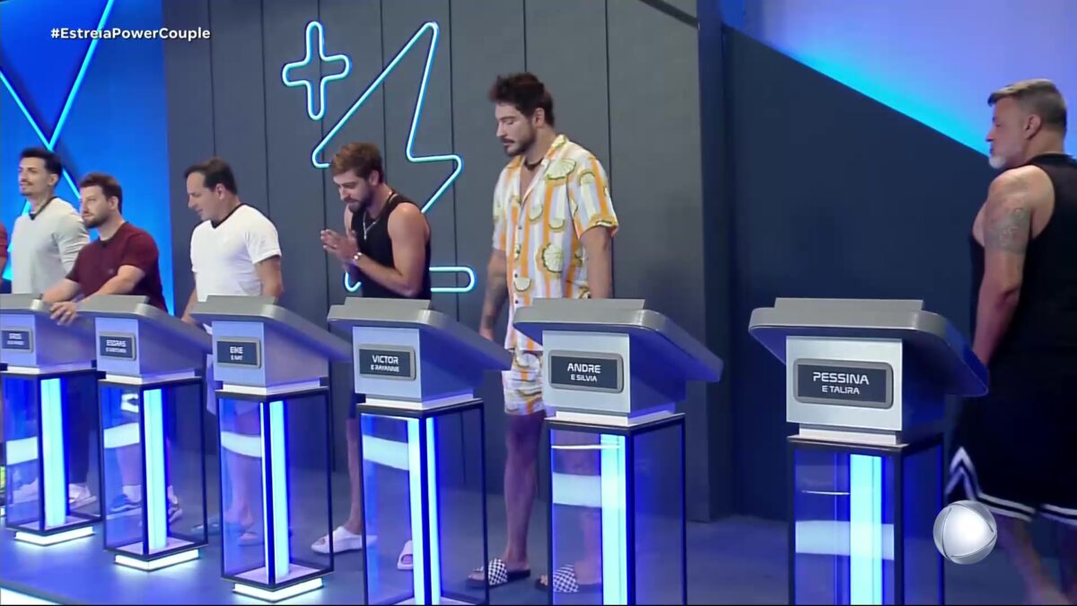 Homens na sala de apostas do Power Couple Brasil 7. Eles estão posicionados atrás de púlpitos com seus respectivos nomes