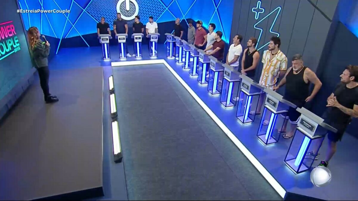 Homens posicionados em seus púlpitos na sala de apostas da 1ª Prova das Mulheres do Power Couple Brasil 7. Rafa Brites está no palco apresentando