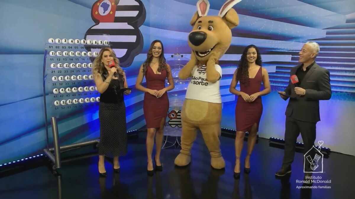 Mascote do São Paulo dá Sorte com orelhas de coelho ao lado das assistentes de palco e dos apresentadores no sorteio deste domingo 20 de abril