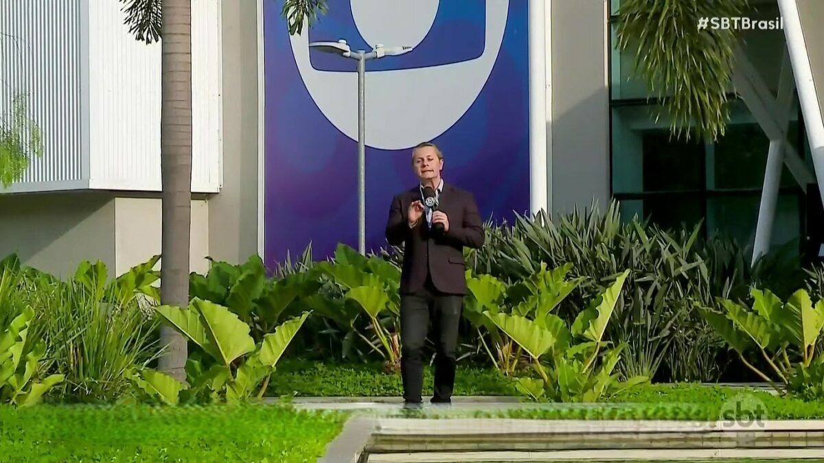 repórter Léo Sant’Anna gravando reportagem do SBT Brasil no jardim dos Estúdios Globo, em frente ao logo da TV Globo em azul