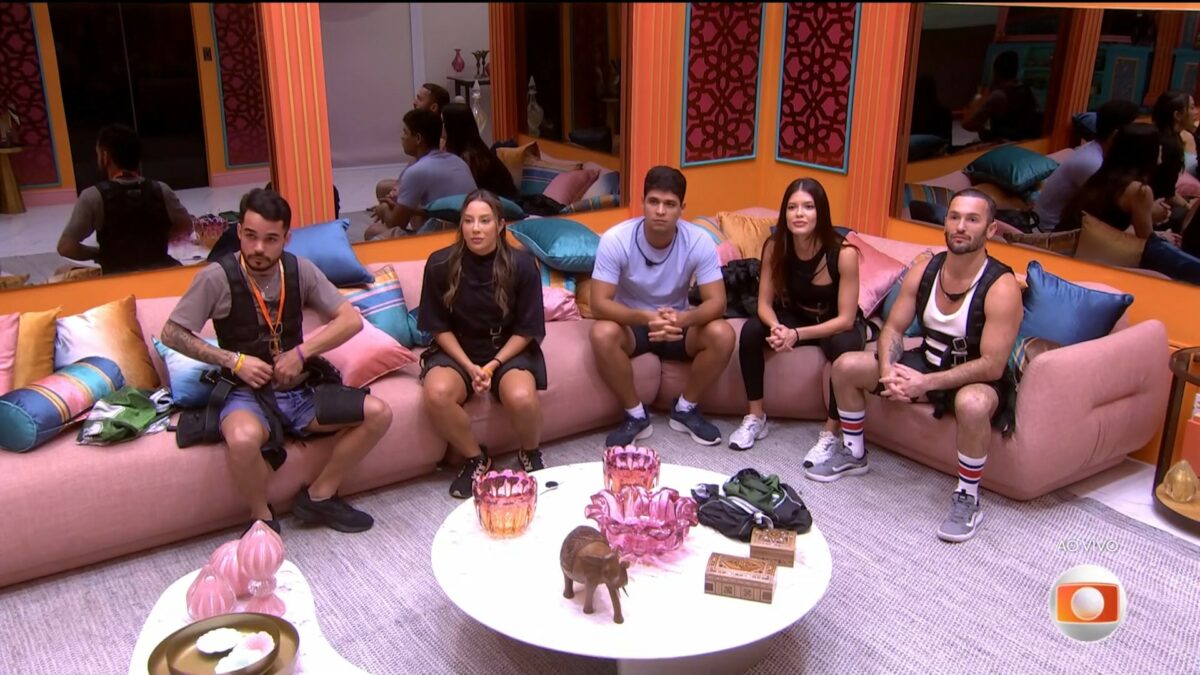 João Pedro, Renata, Guilherme, Vitória Strada e Diego Hypolito sentados no sofá da sala do BBB 25 olhando para o telão e com roupa para disputar prova
