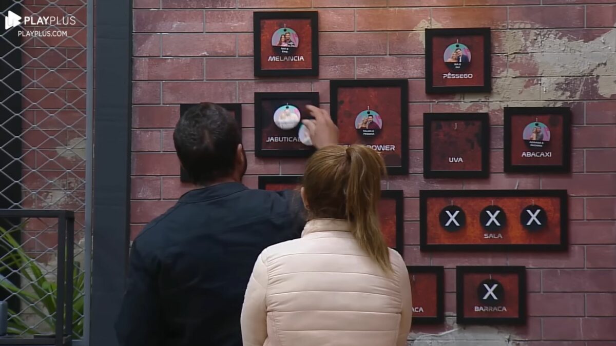 Adriana e Dhomini em frente ao quadro da divisão de quartos no Power Couple Brasil 7