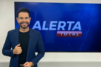 Apresentador Edinho Fernandes sorridente, em frente ao telão do Alerta Total