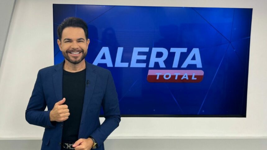 Apresentador Edinho Fernandes sorridente, em frente ao telão do Alerta Total