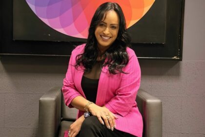 Lisa Gomes sentada em uma poltrona, em frente ao logo do SBT