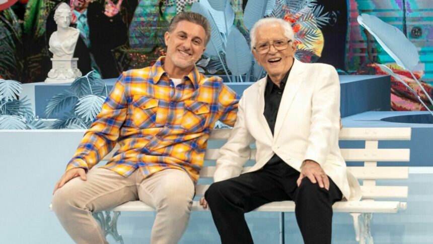 Luciano Huck e Carlos Alberto de Nóbrega sentados em um banco de praça no Domingão