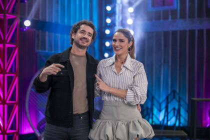 Felipe Andreoli sorrindo enquanto Rafa Brites aponta para ele, também sorrindo, em pé no palco do cenário do Power Couple Brasil 7