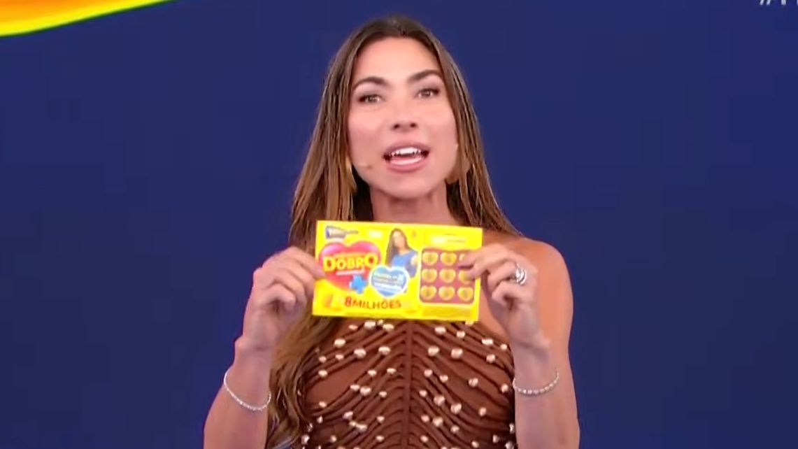 Patricia Abravanel sorrindo e segurando o bilhete do quarto sorteio da Tele Sena de Mães 2025. Ela veste um vestido marrom.