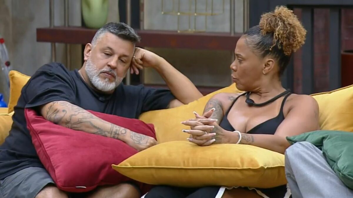 Silvia e André sentados no sofá do Power Couple