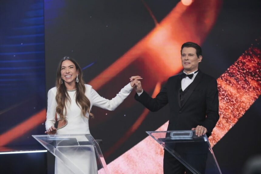 Patricia Abravanel de vestido branco, pega na mão de Celso Portiolli e levanta, na gravação do Troféu Imprensa. Os dois estão sorridentes e Celso veste terno preto com gravata borboleta