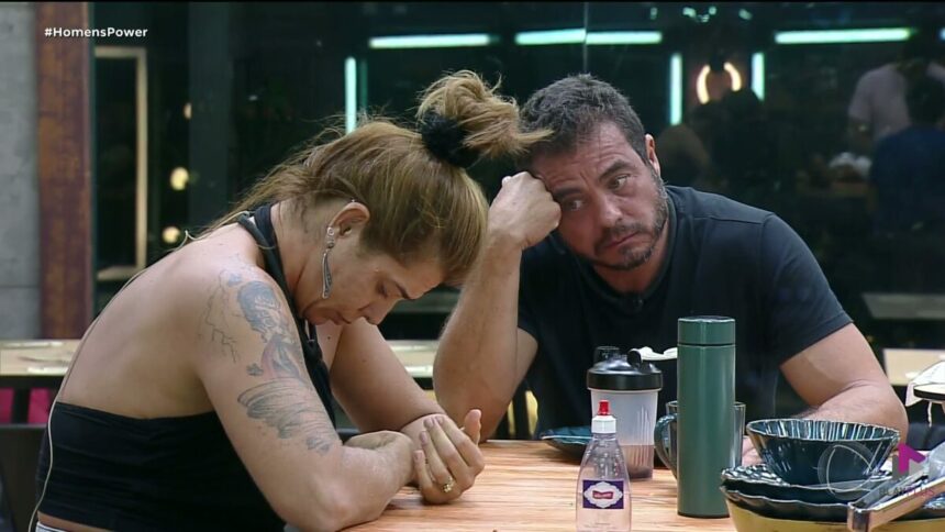Adriana de cabeça baixa sentada na mesa de jantar do Power Couple Brasil, e Dhomini olhando para ela