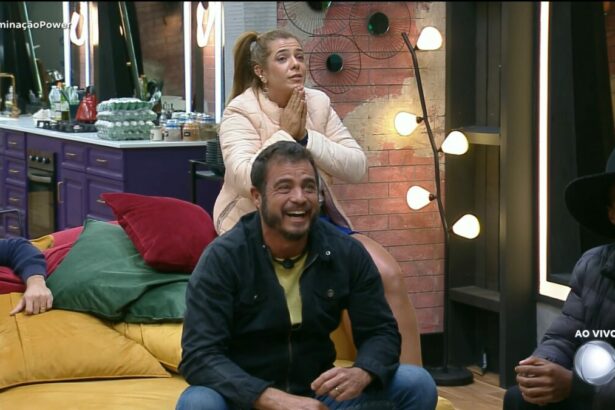 Adriana com as mãos na boca atrás de Dhomini que está sentado no sofá da Mansão do Power Couple Brasil 7