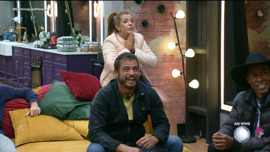 Adriana com as mãos na boca atrás de Dhomini que está sentado no sofá da Mansão do Power Couple Brasil 7