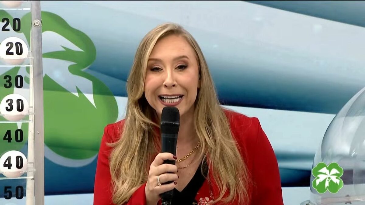 Apresentadora do Alagoas da Sorte em frente ao painel com as bolas do sorteio de 11 de maio. Ela segura um microfone preto