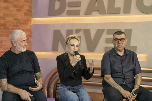 Ana Maria Braga veste blusa preta ao lado dos chefes na coletiva de imprensa do Chef de Alto Nível. Ela segura um microfone da Globo