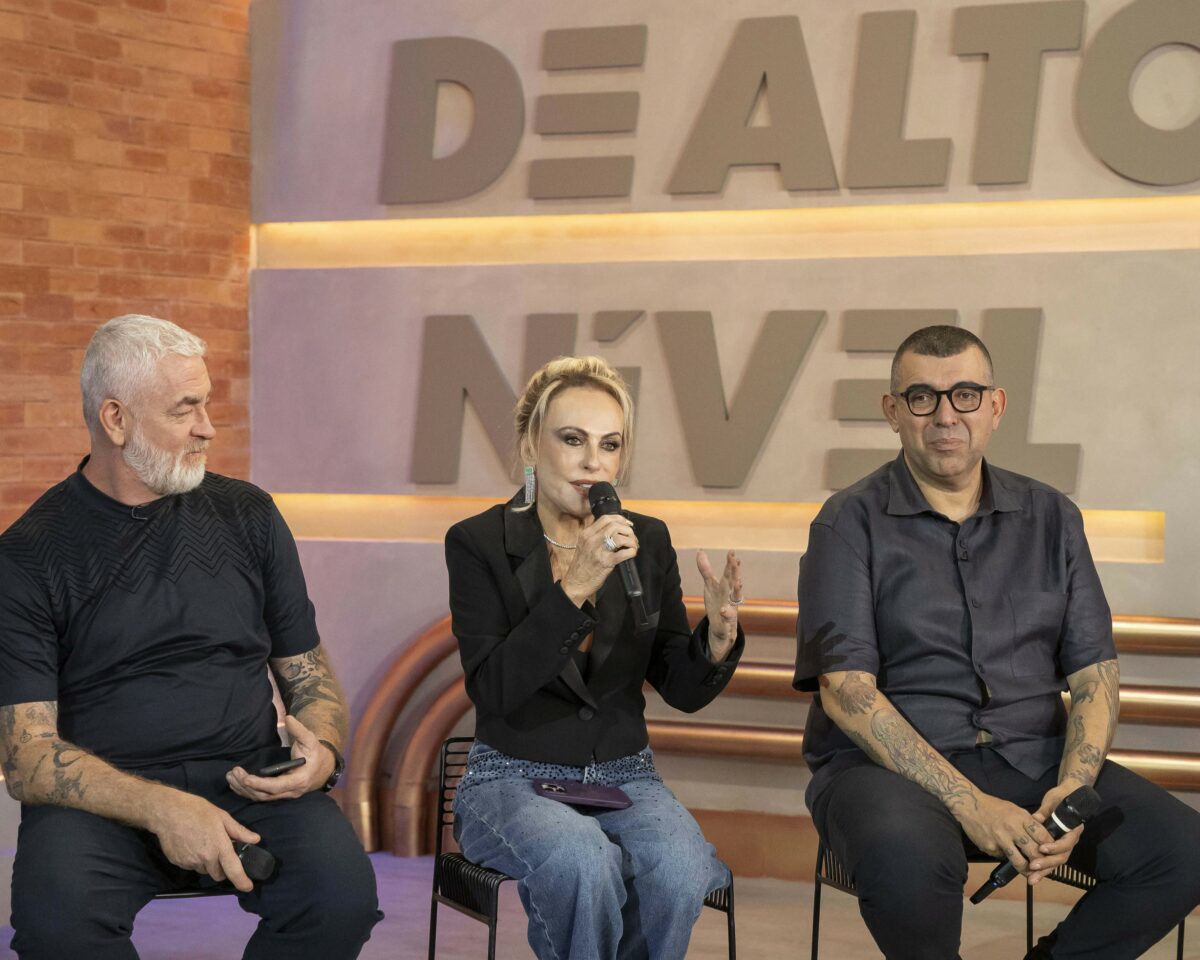 Ana Maria Braga veste blusa preta ao lado dos chefes na coletiva de imprensa do Chef de Alto Nível. Ela segura um microfone da Globo