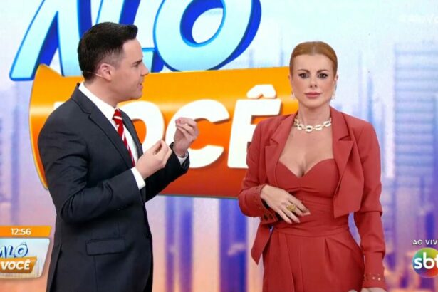 Luiz Bacci gesticula com as mãos e olha para Deborah Albuquerque que olha para frente no cenário do Alô Você