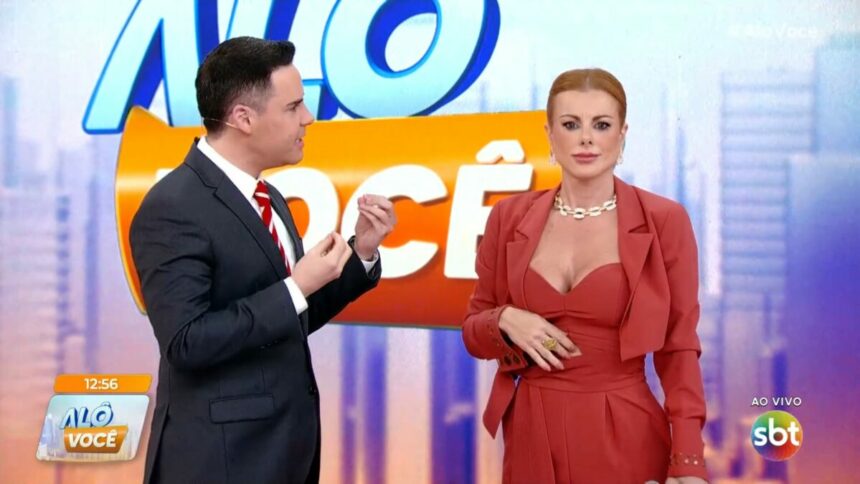 Luiz Bacci gesticula com as mãos e olha para Deborah Albuquerque que olha para frente no cenário do Alô Você