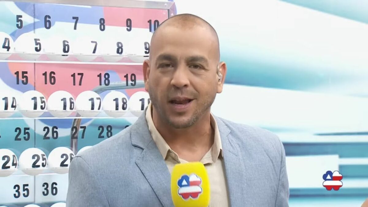 Apresentador do Bahia dá Sorte em frente ao painel com as bolas do sorteio de 11 de maio. Ele segura um microfone amarelo