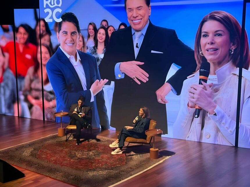 Cesar Filho sentado ao lado de Daniela Beyruti. Eles estão em frente a um telão gigante que trás imagens dos dois e de Silvio Santos no Rio2C