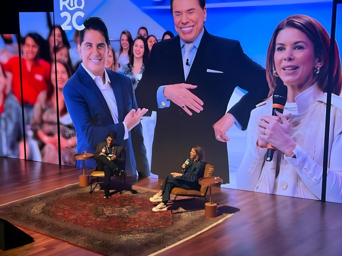 Cesar Filho sentado ao lado de Daniela Beyruti. Eles estão em frente a um telão gigante que trás imagens dos dois e de Silvio Santos no Rio2C
