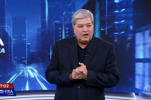 Datena de terno e camisa preta apresentando o Tá na Hora. Ele bate palmas no cenário do SBT
