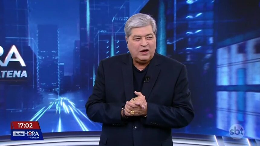 Datena de terno e camisa preta apresentando o Tá na Hora. Ele bate palmas no cenário do SBT