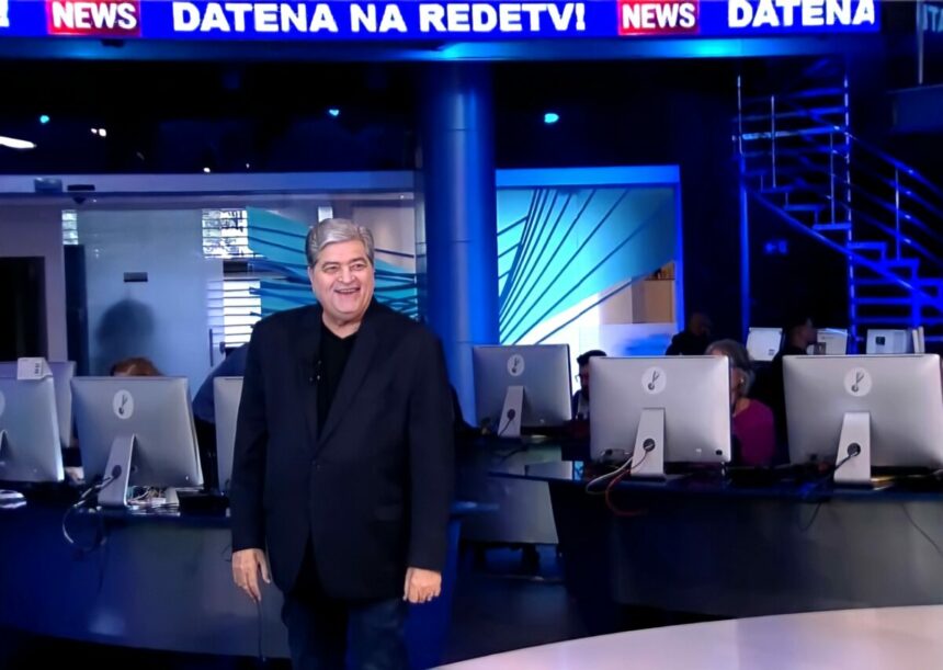 Datena em pé na redação da RedeTV!. Ele está sorridente