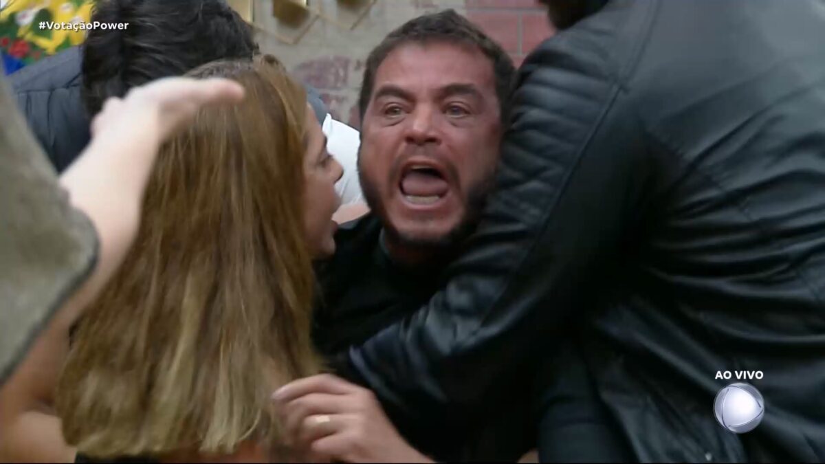 Dhomini gritando contra Carol e sendo segurado por outros participantes no sofá do Power Couple