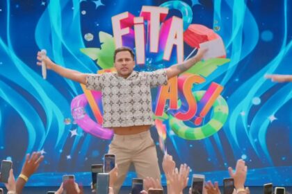 Lucas Guimarães abre os braços em frente ao telão do programa Eita,Lucas! no SBT. Ele está com semblante assustado e segura um microfone branco em uma das mãos