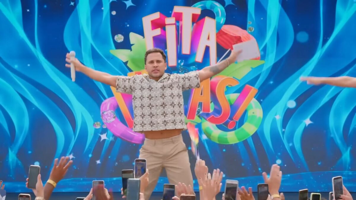 Lucas Guimarães abre os braços em frente ao telão do programa Eita,Lucas! no SBT. Ele está com semblante assustado e segura um microfone branco em uma das mãos