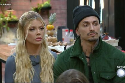 Giovanna e Diego olham assustados para os demais participantes na sala do Power Couple Brasil 7. Eles estão sentados em um banquinho da DR