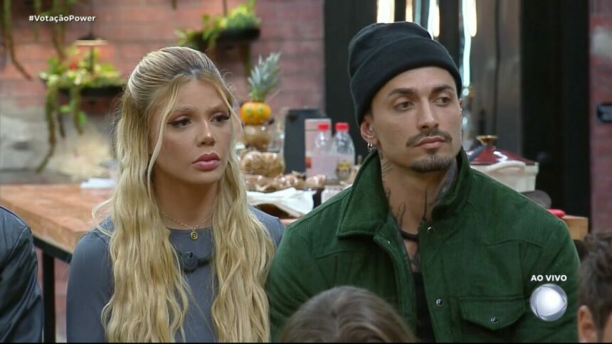 Giovanna e Diego olham assustados para os demais participantes na sala do Power Couple Brasil 7. Eles estão sentados em um banquinho da DR