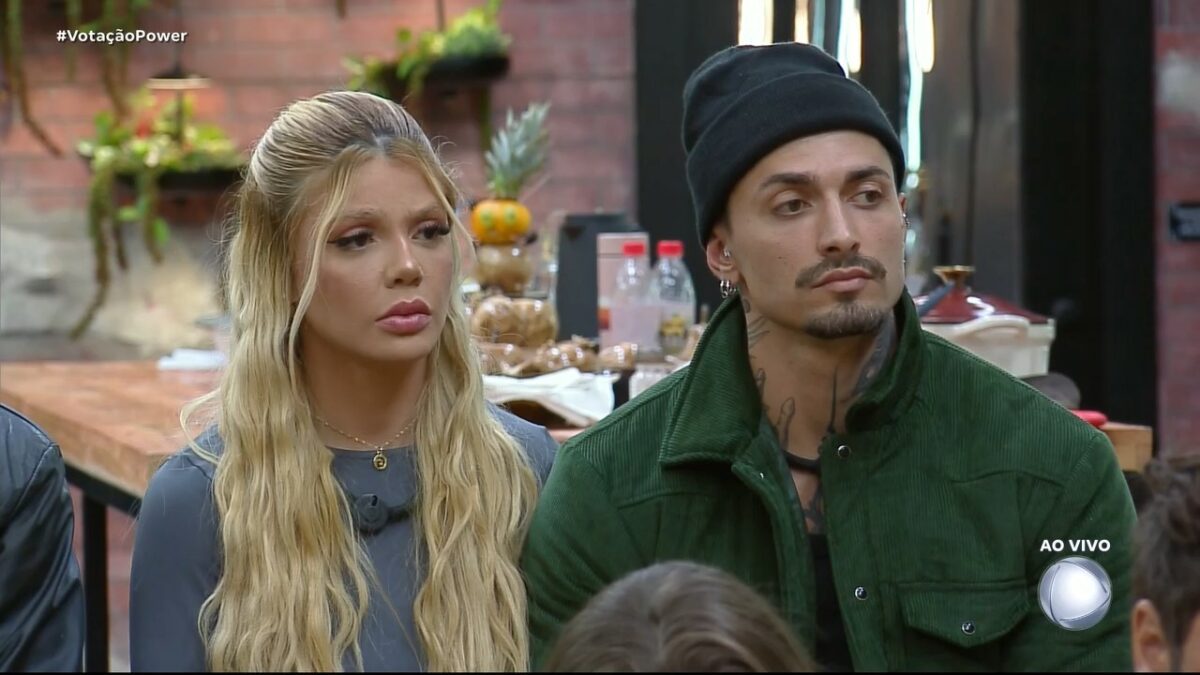 Giovanna e Diego olham assustados para os demais participantes na sala do Power Couple Brasil 7. Eles estão sentados em um banquinho da DR