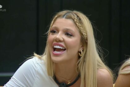 Giovanna de blusa branca, sentada no sofá da Mansão Power, sorrindo ao ver Diego que disputava a Prova dos Homens
