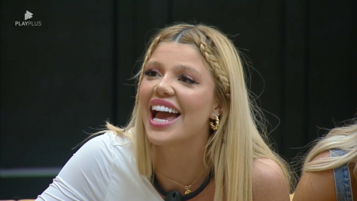 Giovanna de blusa branca, sentada no sofá da Mansão Power, sorrindo ao ver Diego que disputava a Prova dos Homens