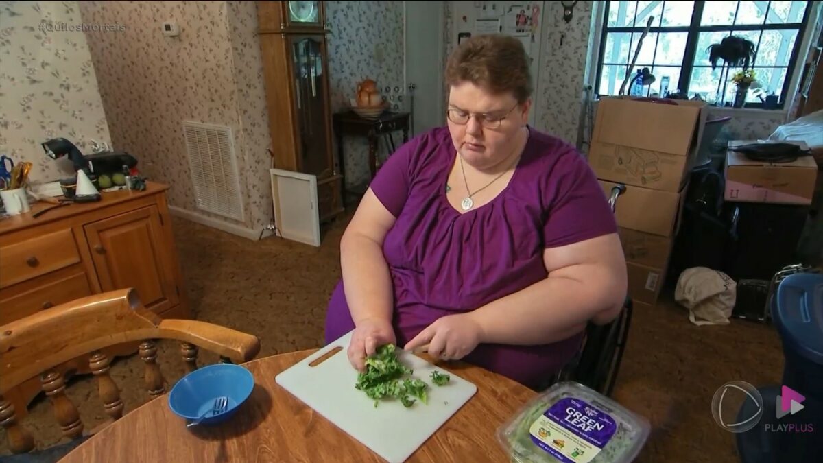 Holly, de blusa vinho, sentada em uma cadeira de rodas cortando alfaces em cena do Quilos Mortais