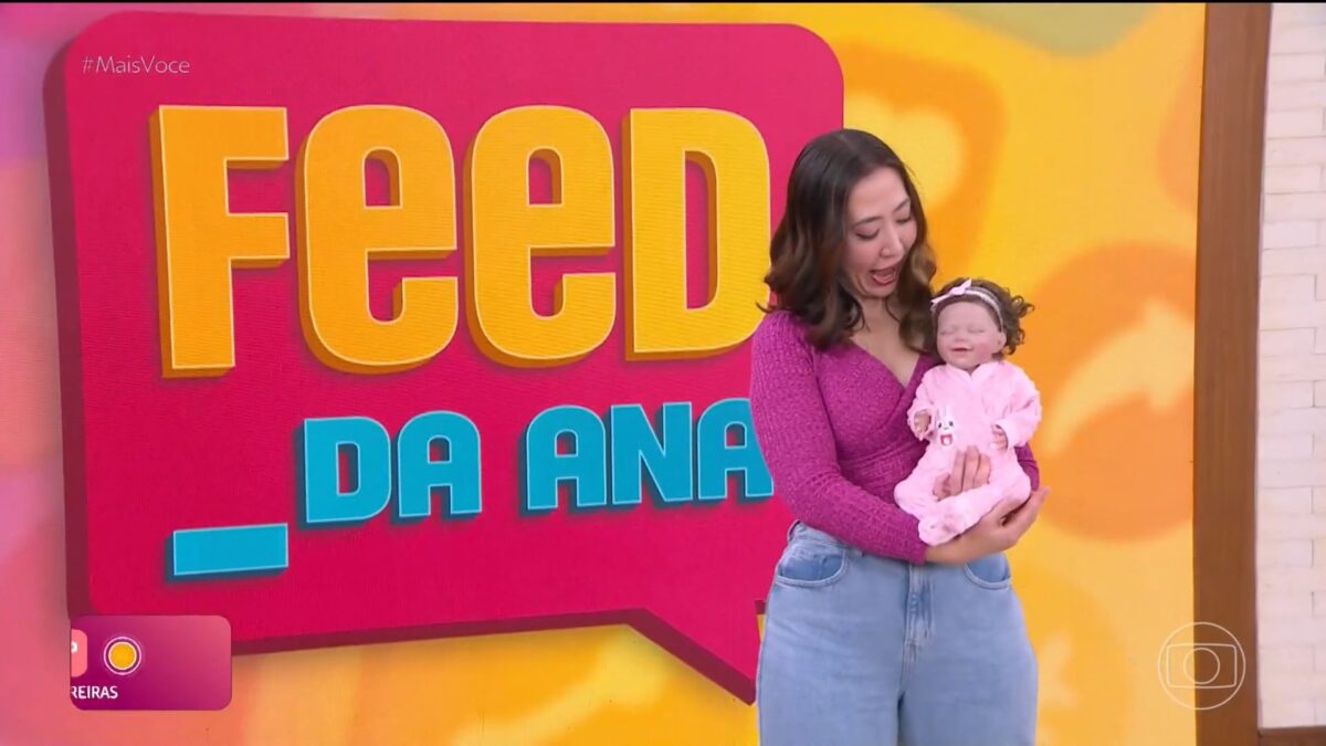 Juju Massaoka segura Bebê Reborn em frente ao telão do Feed da Ana do Mais Você. A repórter olha o bebê com a boca aberta