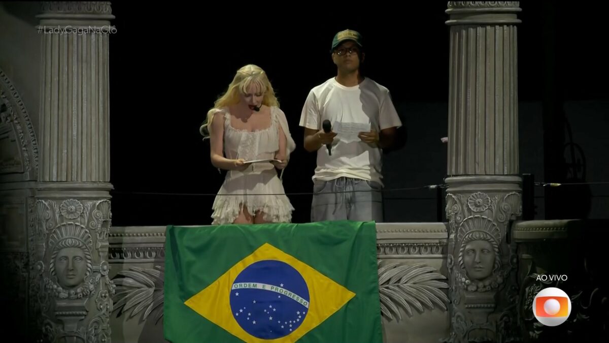 Lady Gaga olha para carta que leu para o Brasil ao lado do intérprete em show. Ela está em pé numa janela do cenário do palco com a bandeira do país asteada
