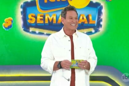 Luis Ricardo veste camisa branca, segura o bilhete da Tele Sena Semana edição 50 e está sorrindo. O cenário está verde e e ele se posiciona a frente dos globos com as bolas do sorteio