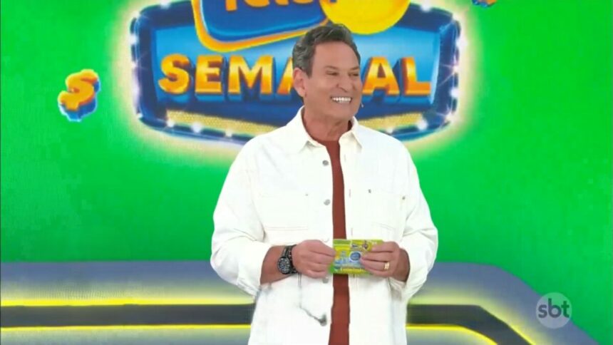 Luis Ricardo veste camisa branca, segura o bilhete da Tele Sena Semana edição 50 e está sorrindo. O cenário está verde e e ele se posiciona a frente dos globos com as bolas do sorteio