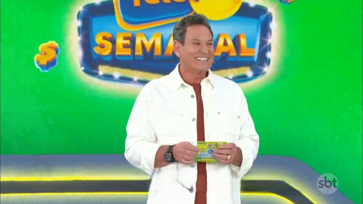 Luis Ricardo veste camisa branca, segura o bilhete da Tele Sena Semana edição 50 e está sorrindo. O cenário está verde e e ele se posiciona a frente dos globos com as bolas do sorteio